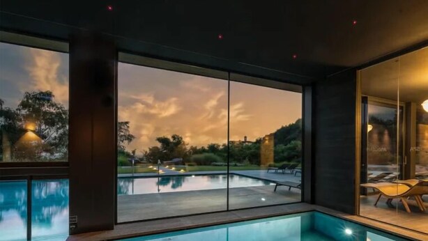 Una piscina interior moderna amb grans portes de vidre que donen a una vista exterior panoràmica durant el capvespre.
