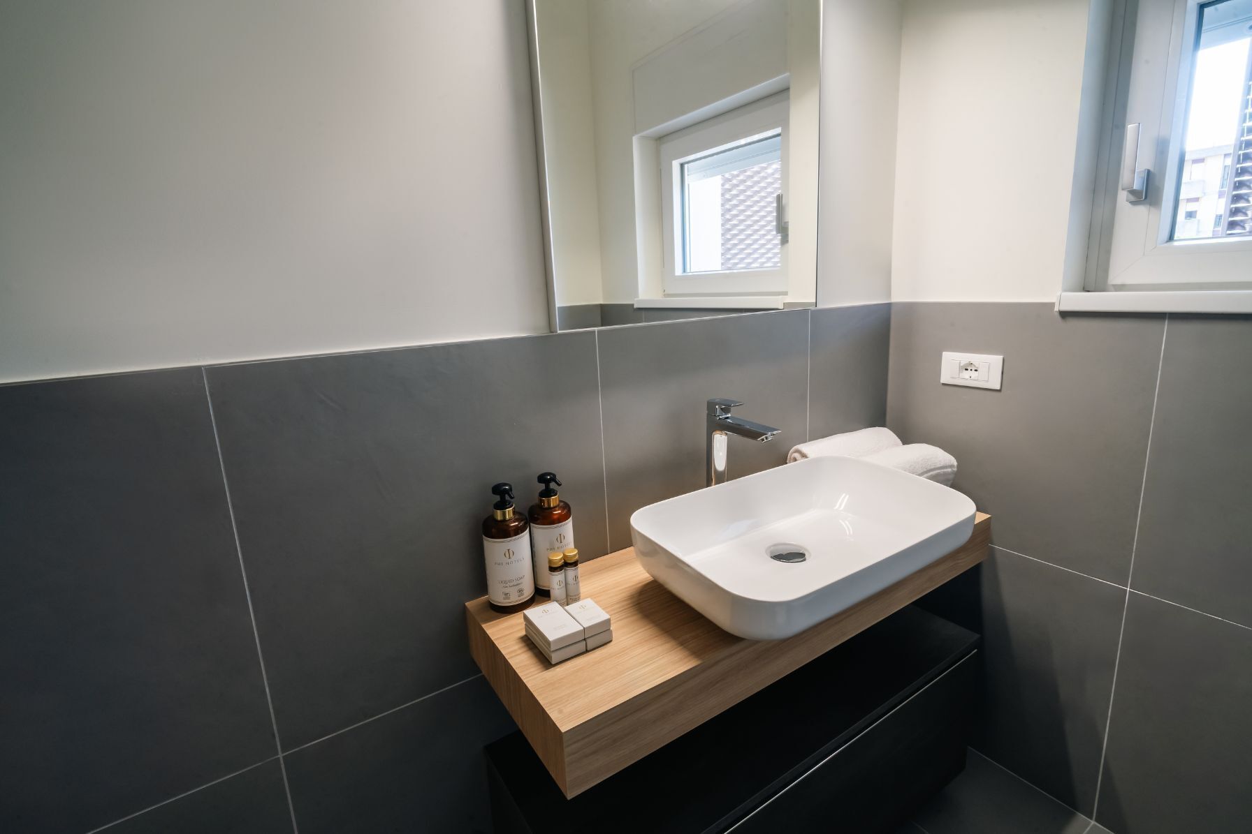 A modern bathroom with a white sink, dark tiles, a wooden vanity, and a window above the toilet.