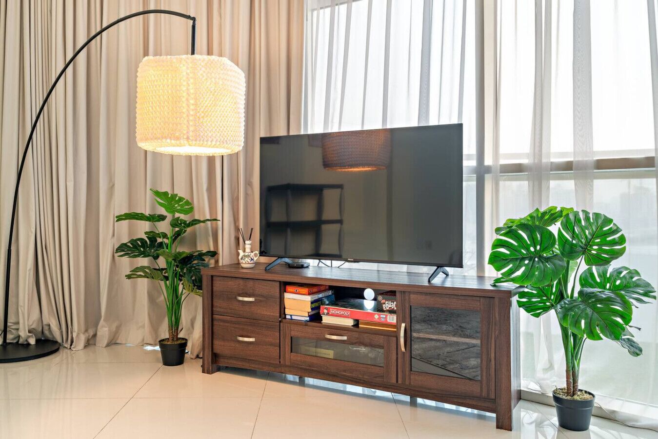 A modern living room with a flat-screen TV on a wooden cabinet, two potted plants, a floor lamp, and a large window with sheer curtains.
