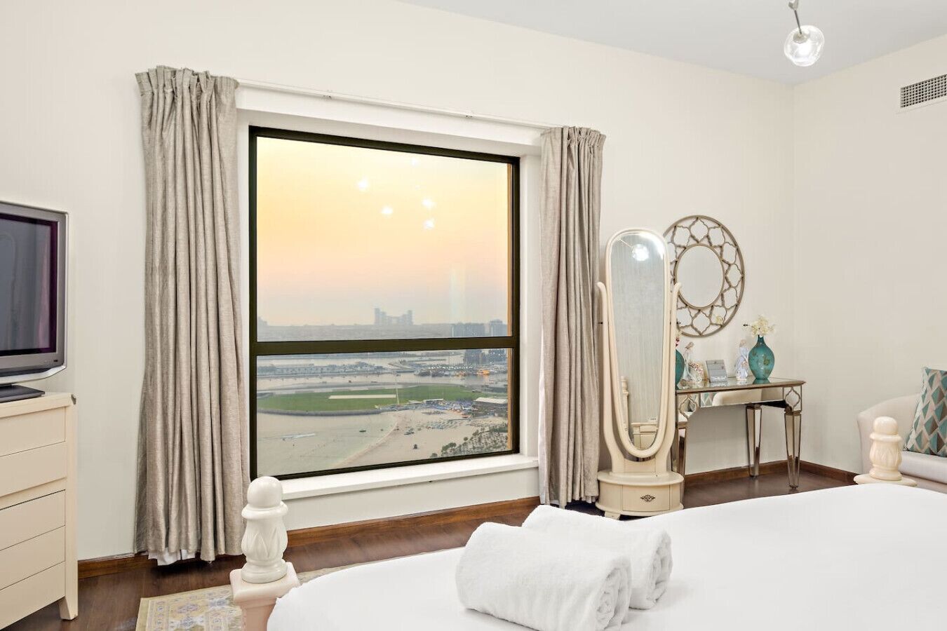 A hotel room with a large window, a bed, a TV, a vanity with a mirror, and a desk with a vase.