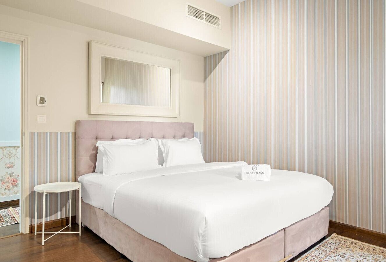A neatly made bed with a headboard, a small bedside table, and a framed mirror on the wall.