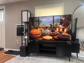A flat-screen TV on a black stand with a scenic image on the screen, a speaker, and a window with blinds.