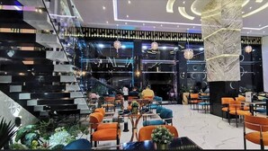 Un moderno vestíbulo de hotel con una columna central de mármol, varias zonas de descanso y una escalera que conduce a pisos superiores.