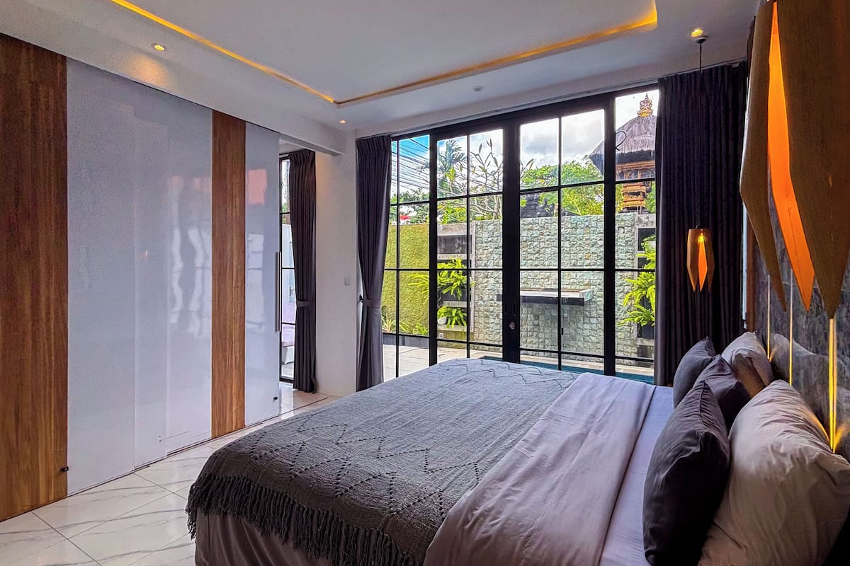 A modern bedroom with a large bed, a view of greenery and a building outside the window, and a large window with curtains.