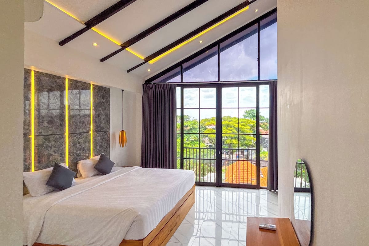 A modern bedroom with a large bed, a view of greenery outside, and a balcony with a railing.