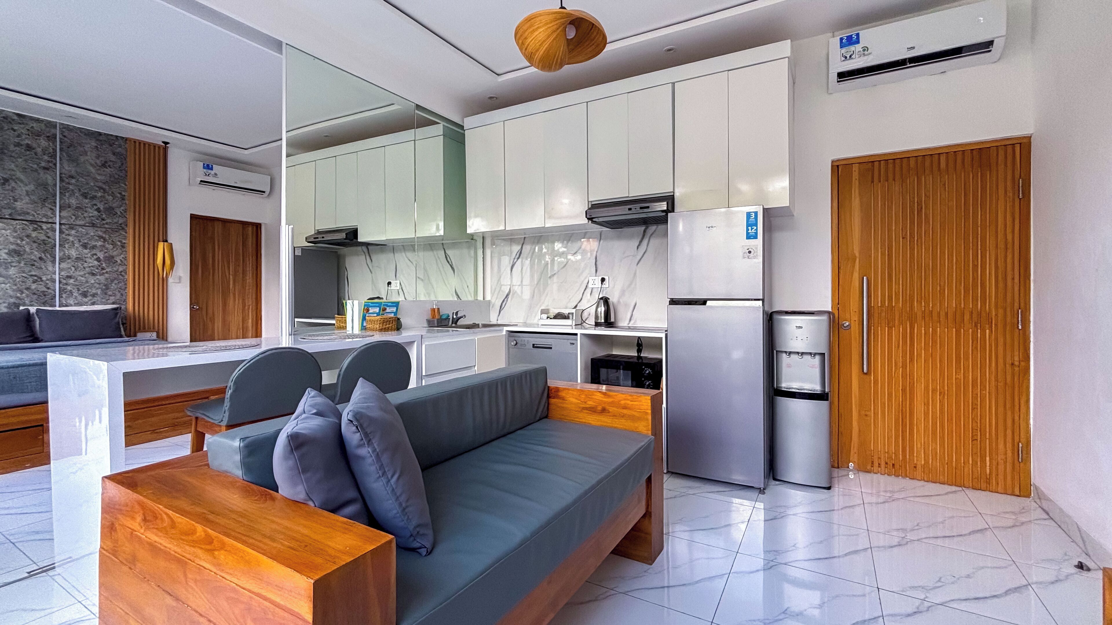 A modern kitchen with white cabinets, a built-in oven, and a refrigerator. There is a wooden door and a small dining area with a bench and cushions.