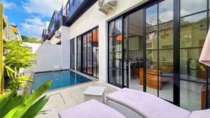 A modern outdoor pool area with a glass-enclosed patio, lounge seating, and a view of the adjacent building. - Seminyak Loft 360 Bali (Seminyak)