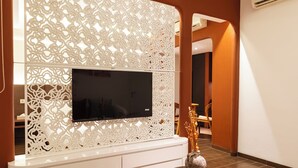 A modern living room with a flat-screen TV mounted on a decorative white panel, a glass coffee table with a decorative vase, and a wooden chair. - Omar Hotel (Tangerang)