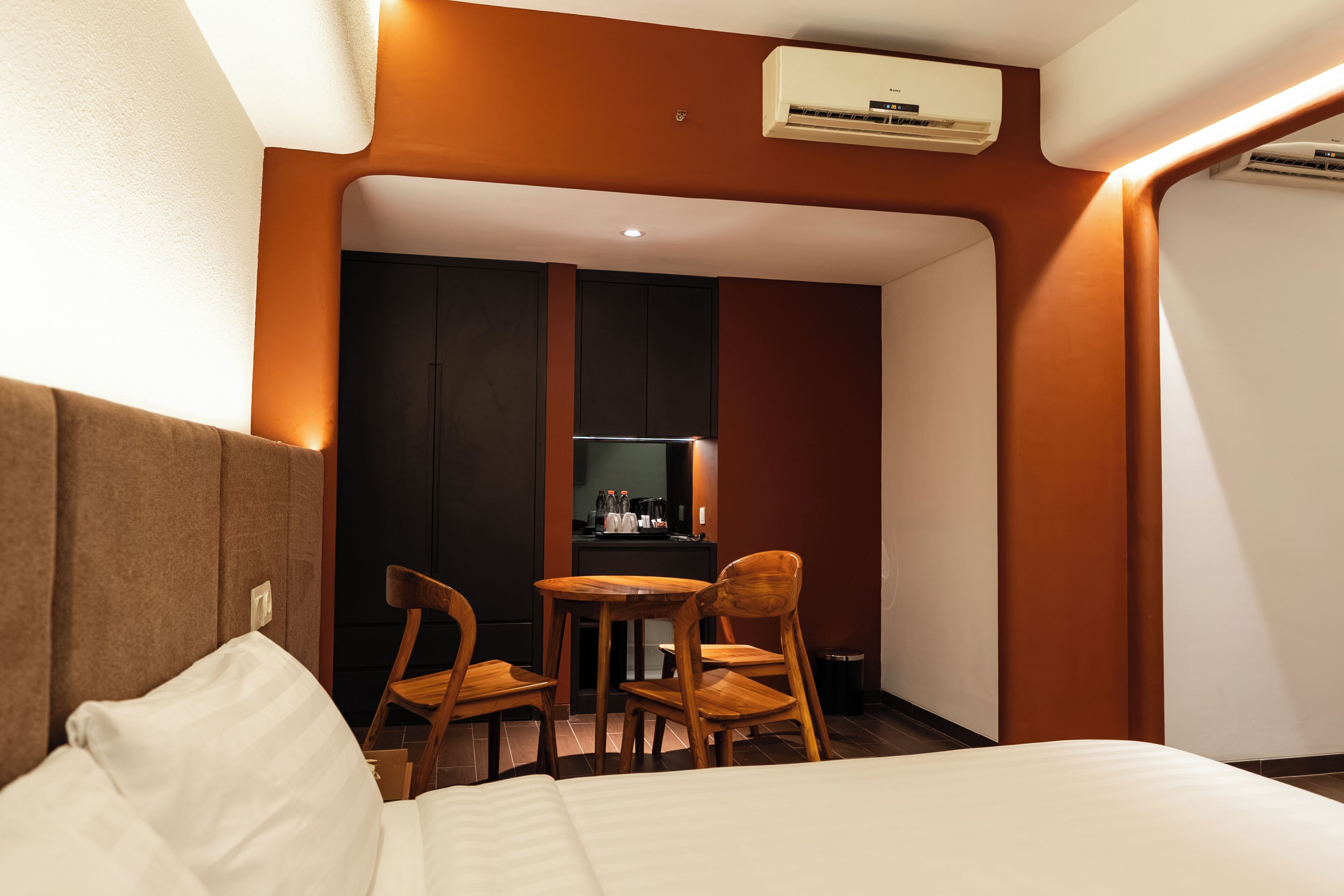 A hotel room with a bed, a small table with chairs, a kitchenette, and an air conditioning unit.