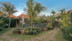 A tropical garden with a stone pathway leading to a building with a red-tiled roof. - Matahari Lumbung Resort (Ubud)