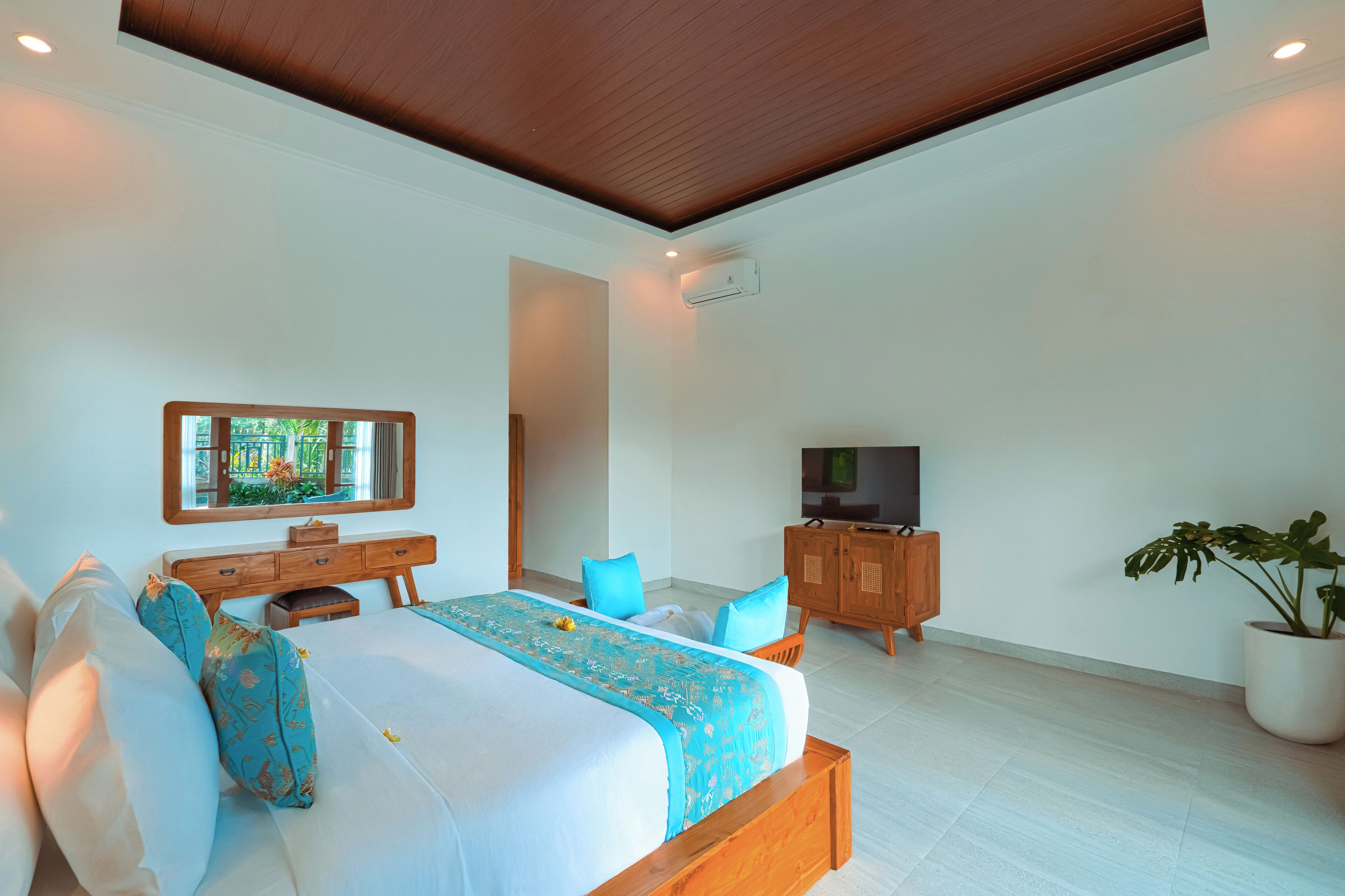 A modern bedroom with a large bed, a wooden headboard, a TV, a desk, and a potted plant.