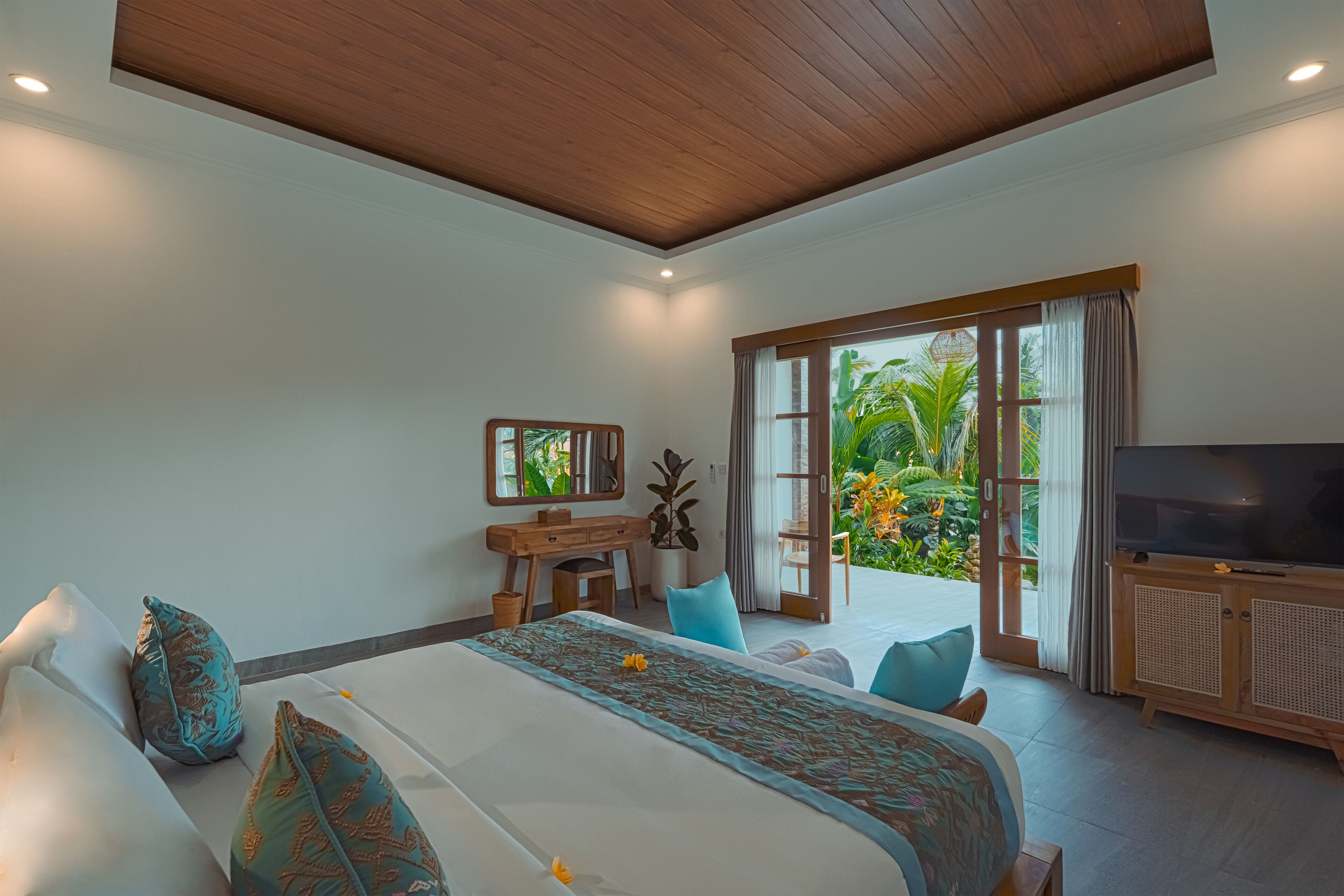 A spacious bedroom with a large bed, a wooden desk, a television, and a sliding glass door leading to a balcony with a view of greenery.