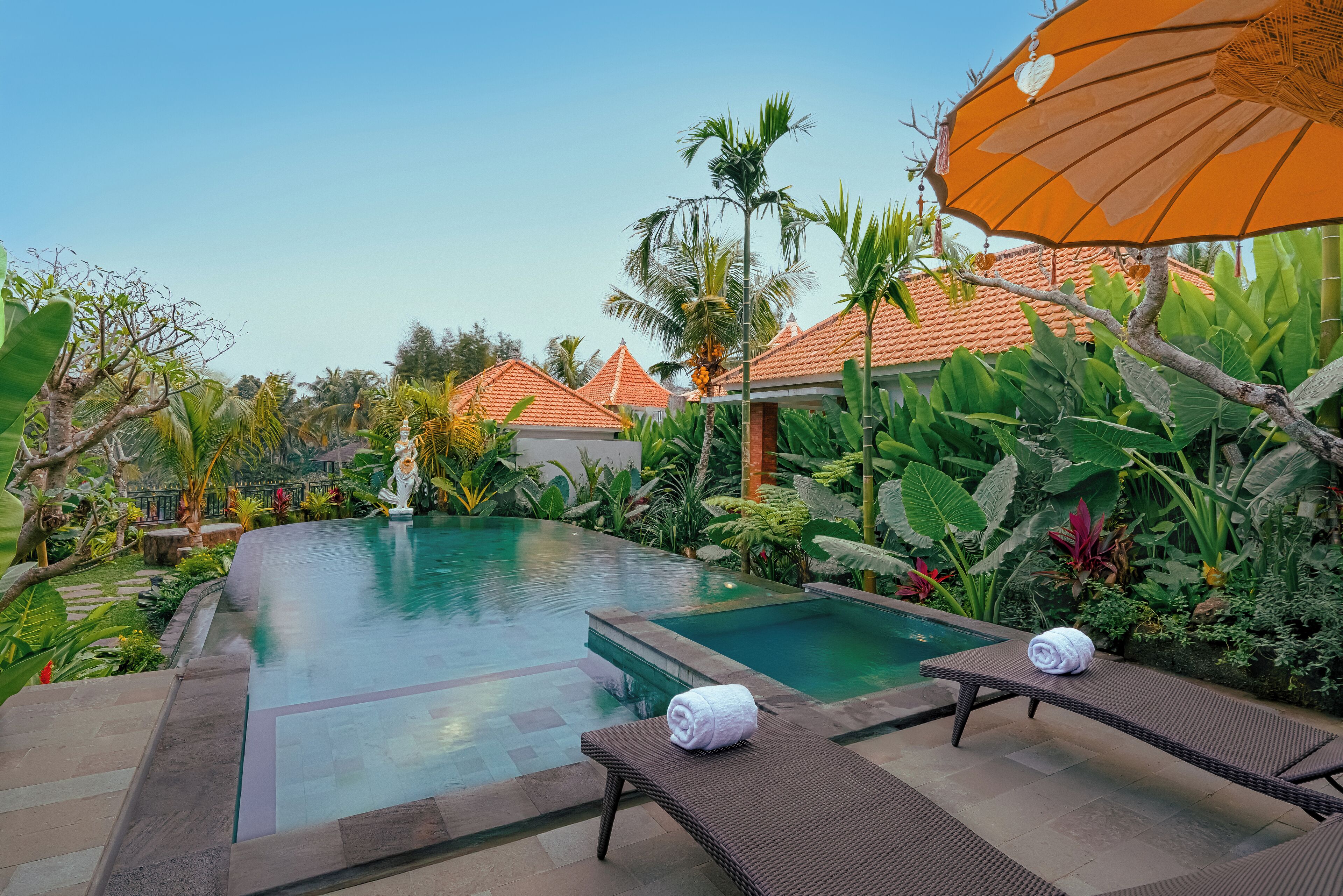 A swimming pool with lounge chairs and an umbrella, surrounded by tropical plants and a clear sky.