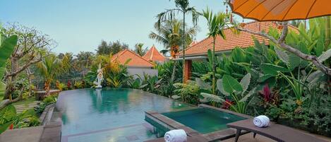 A swimming pool with lounge chairs and an umbrella, surrounded by tropical plants and a clear sky.