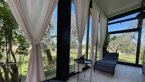 Una habitación con ventanas amplias, un sillón negro, una mesita con una botella y follaje verde en el exterior.