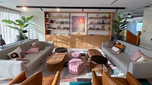 A modern living room with two sofas, a coffee table, and ottomans. There is a bookshelf with various items and a large window providing natural light.
