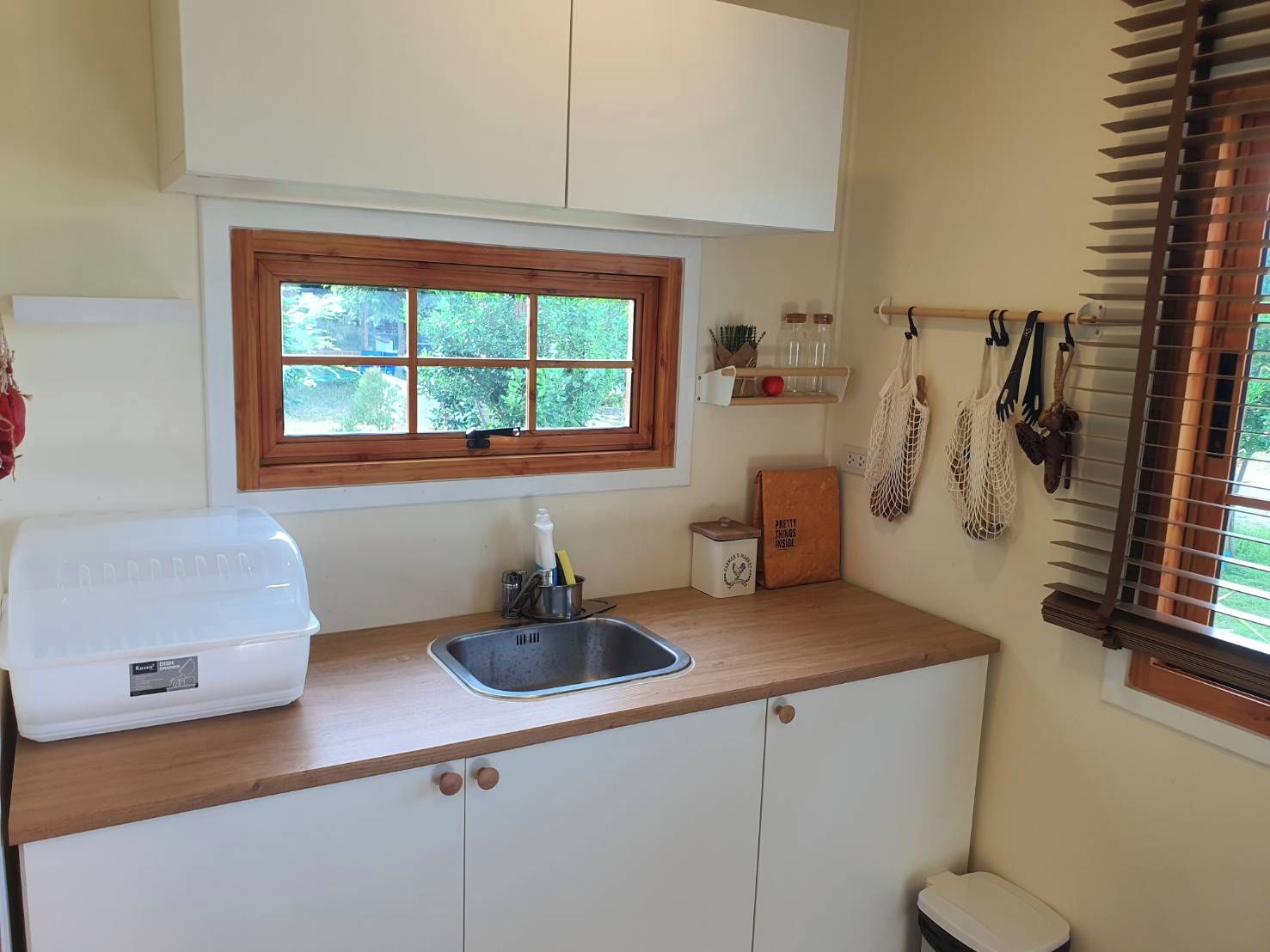 Una cocina con encimera de madera, un fregadero, una ventana con vistas a zonas verdes, y un estante pequeño con frascos y una planta.