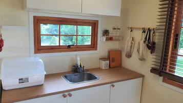 Una cocina con encimera de madera, un fregadero, una ventana con vistas a zonas verdes, y un estante pequeño con frascos y una planta.