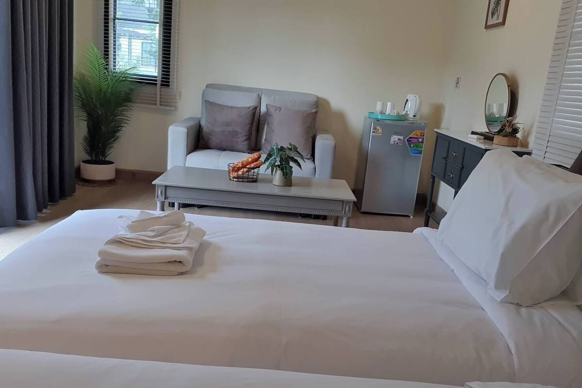 A hotel room with a bed, a sofa, a small table with a plant, a refrigerator, and a window with curtains.