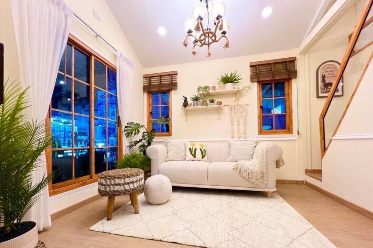 A cozy living room with a white sofa, a round white ottoman, and a geometric rug. There are two windows with wooden frames, a chandelier, and a staircase to the right.