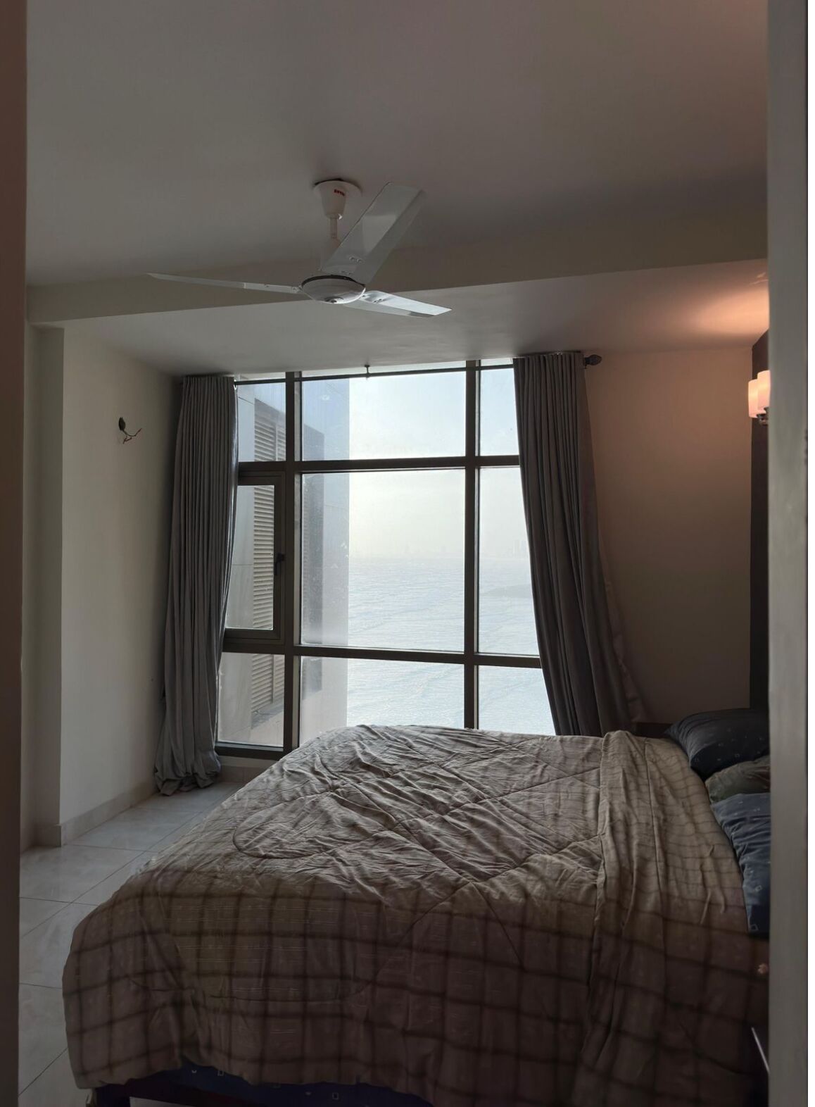 A hotel room with a large window, a bed, a ceiling fan, and a nightstand with a lamp.
