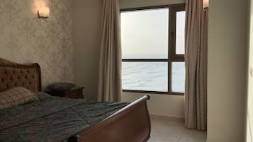 A bedroom with a large bed, a ceiling fan, a nightstand, a window with curtains, and a view of the sea.