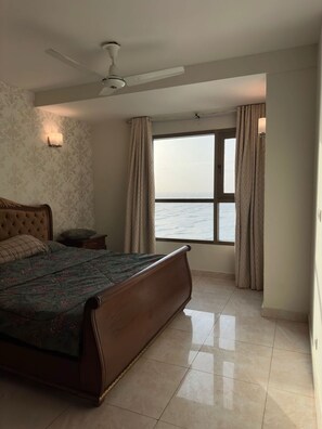 Un dormitorio con una cama grande, un ventilador de techo, una mesita de noche, una ventana con cortinas y vista al mar.