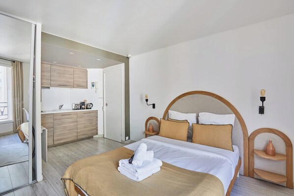 A hotel room with a bed, a wooden headboard, a bedside table, a mirror, and a view of the kitchen and balcony. - Modern Studio Montaigne Champs Elysees (Paris)
