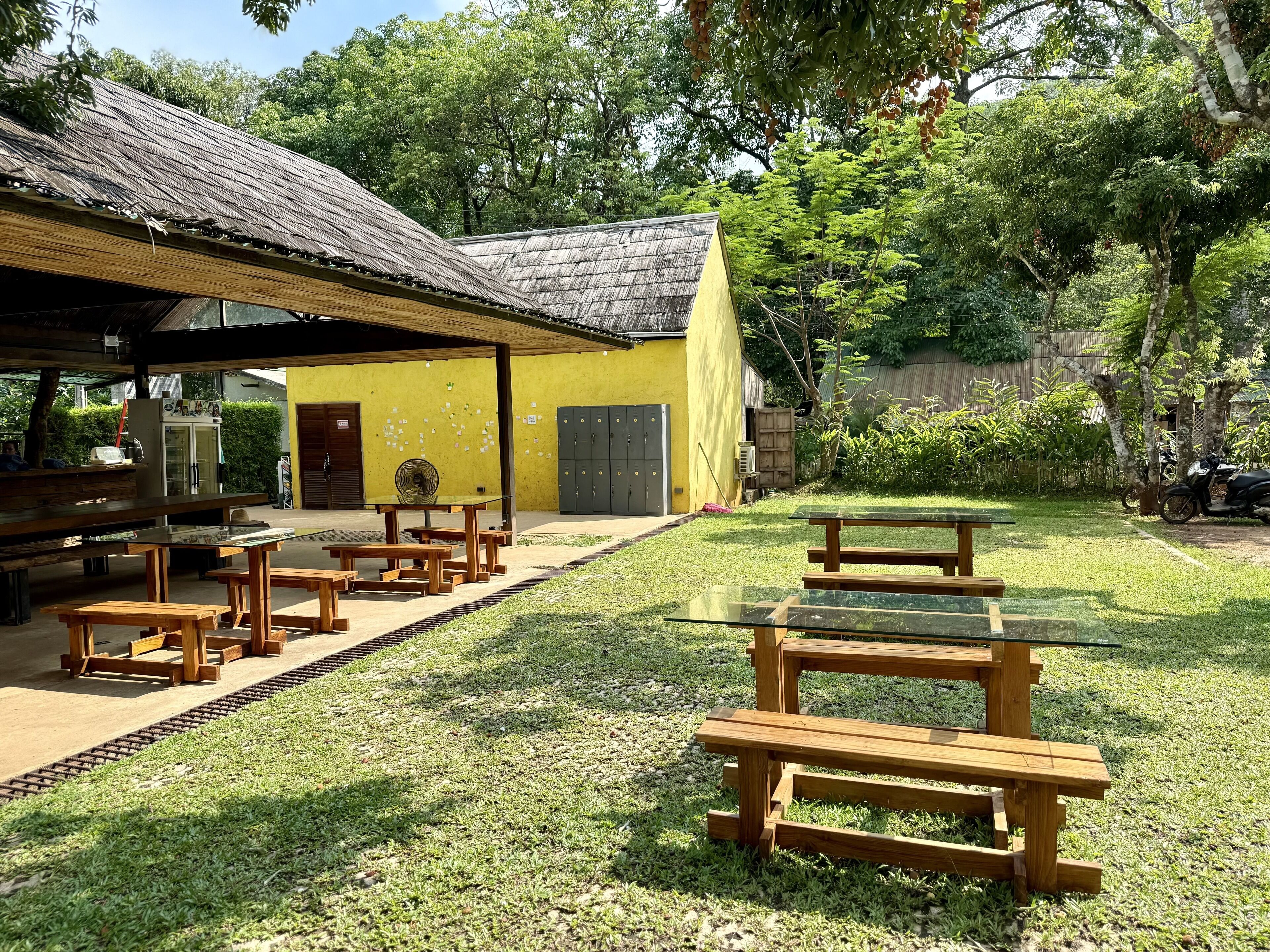 An outdoor dining area with wooden picnic tables and benches, a yellow building with a thatched roof, and a green lawn.