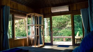 A room with a bed, a wooden bookshelf, and large windows offering a view of greenery.