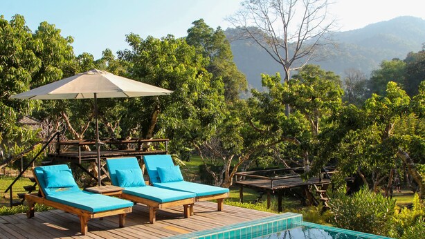 A pool area with two lounge chairs and a canopy, surrounded by lush greenery and a view of distant mountains.