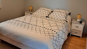 Un lit bien fait, recouvert d’une couvre-lit à motifs géométriques, une table de chevet avec une lampe et une petite table de nuit avec une télécommande.