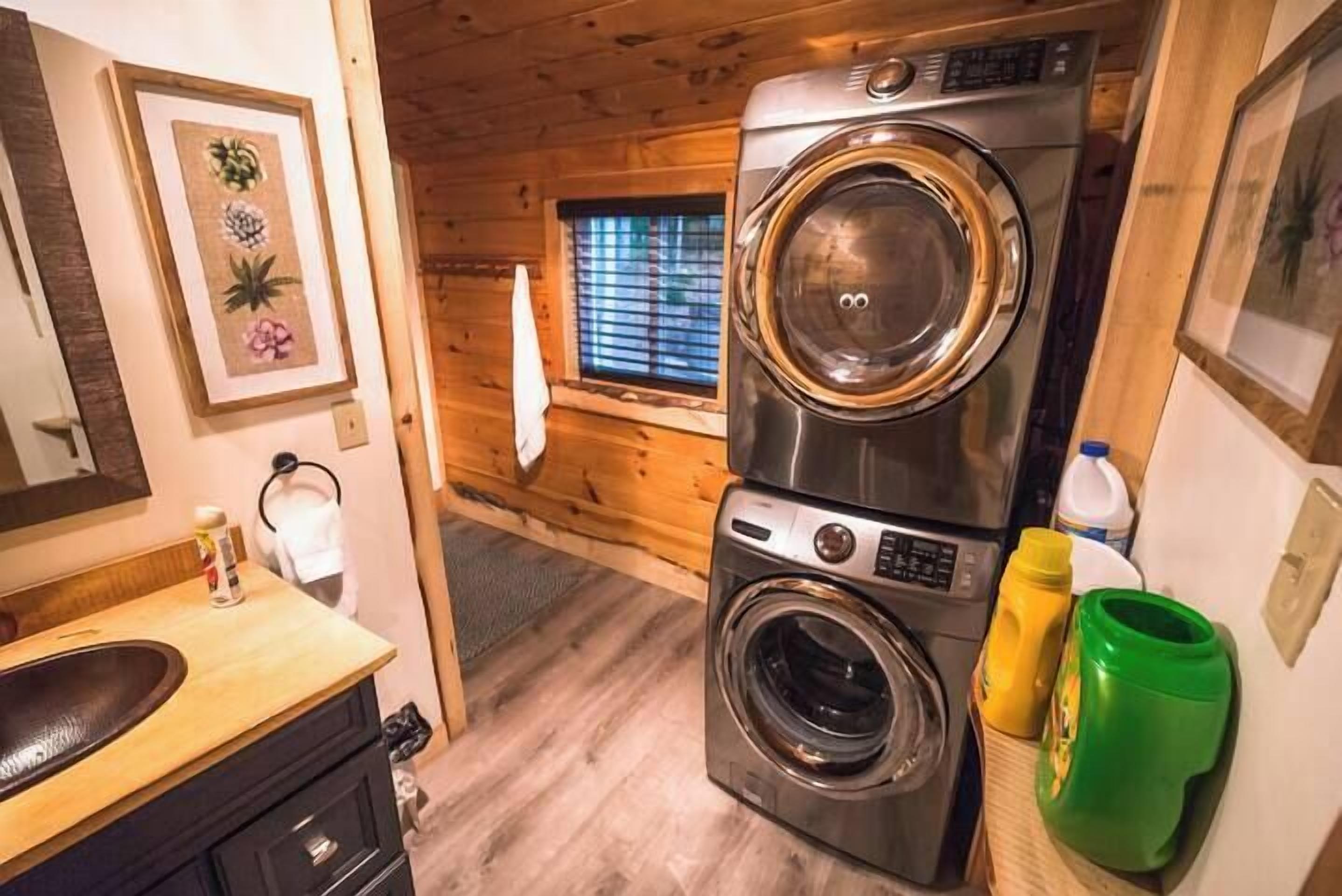 A compact laundry room with a front-loading washer and dryer, a wooden floor, and a window with blinds.