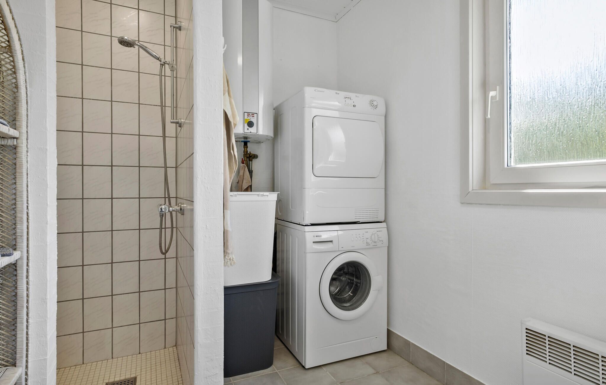 A compact laundry room with a washing machine and dryer stacked on top of each other, a shower with a hose, and a window providing natural light.