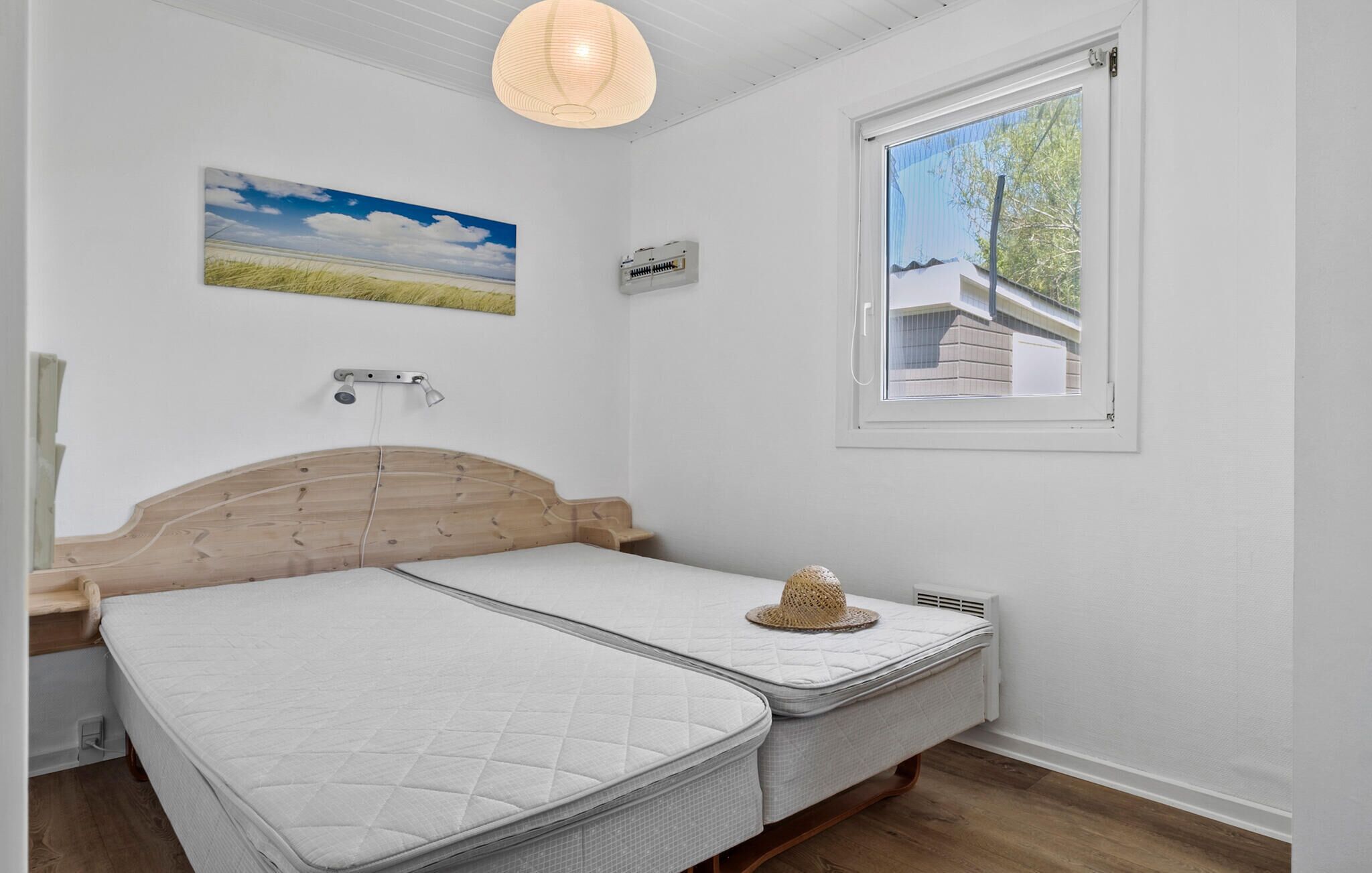 A bedroom with a wooden headboard, a single bed, a small bedside table, a wall-mounted heater, a window with a view of a building, and a ceiling light fixture.