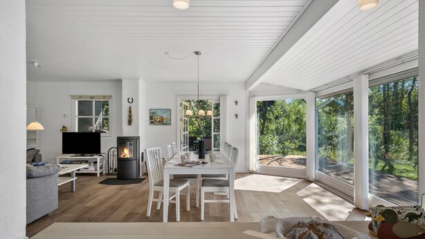 A spacious living area with a dining table, white chairs, a fireplace, and large windows offering a view of the outdoors.