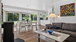A modern living room with a sofa, a dining table with chairs, a kitchen area, and a large window offering a view of the outdoors.