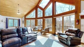 Un vaste salon doté d’un haut plafond et de grandes fenêtres, avec un canapé en cuir, un fauteuil inclinable et une terrasse en bois visible à travers les fenêtres.
