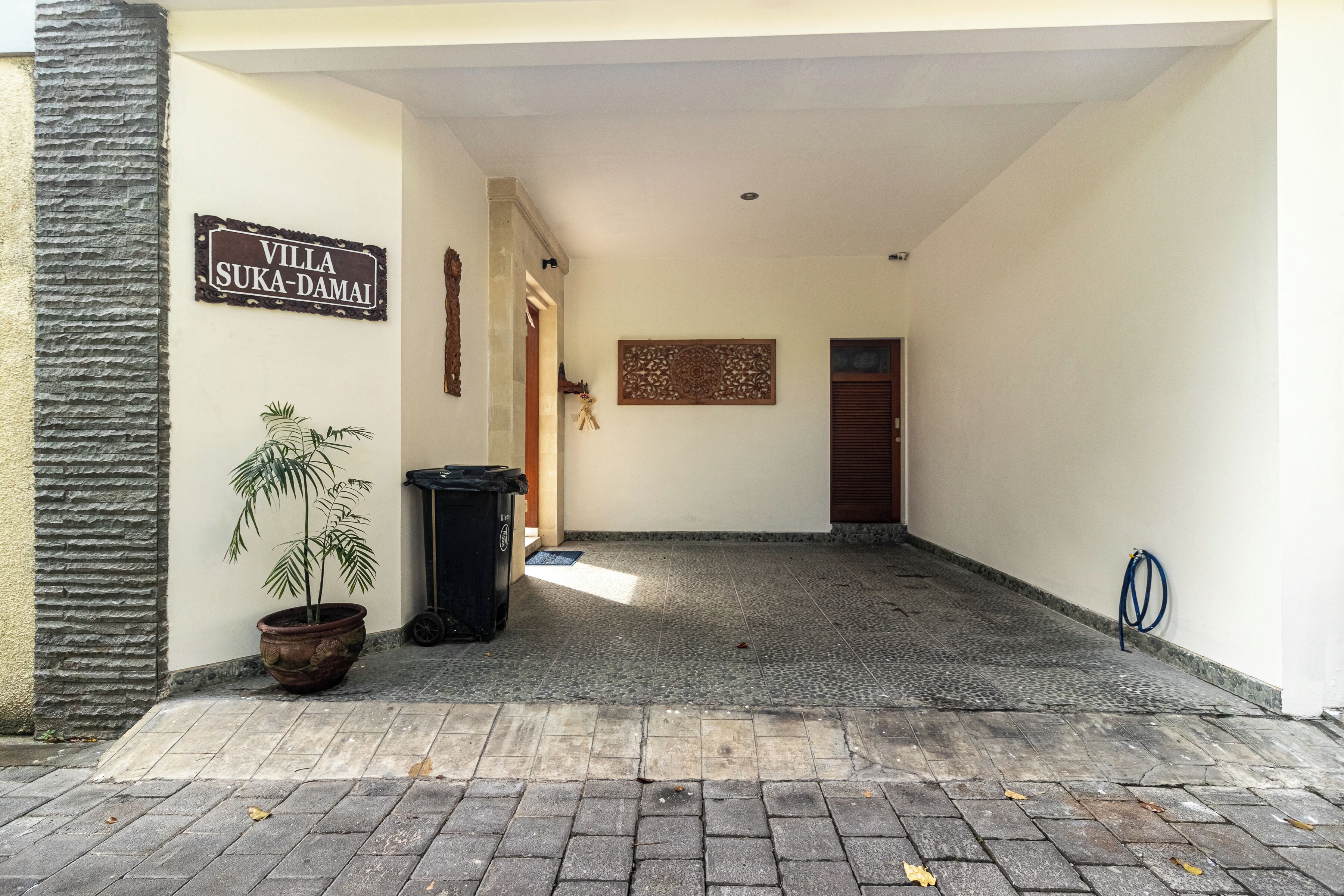 A hallway with a sign reading "Villa Suka-Damai," a potted plant, a trash bin, and a blue hose on the wall.
