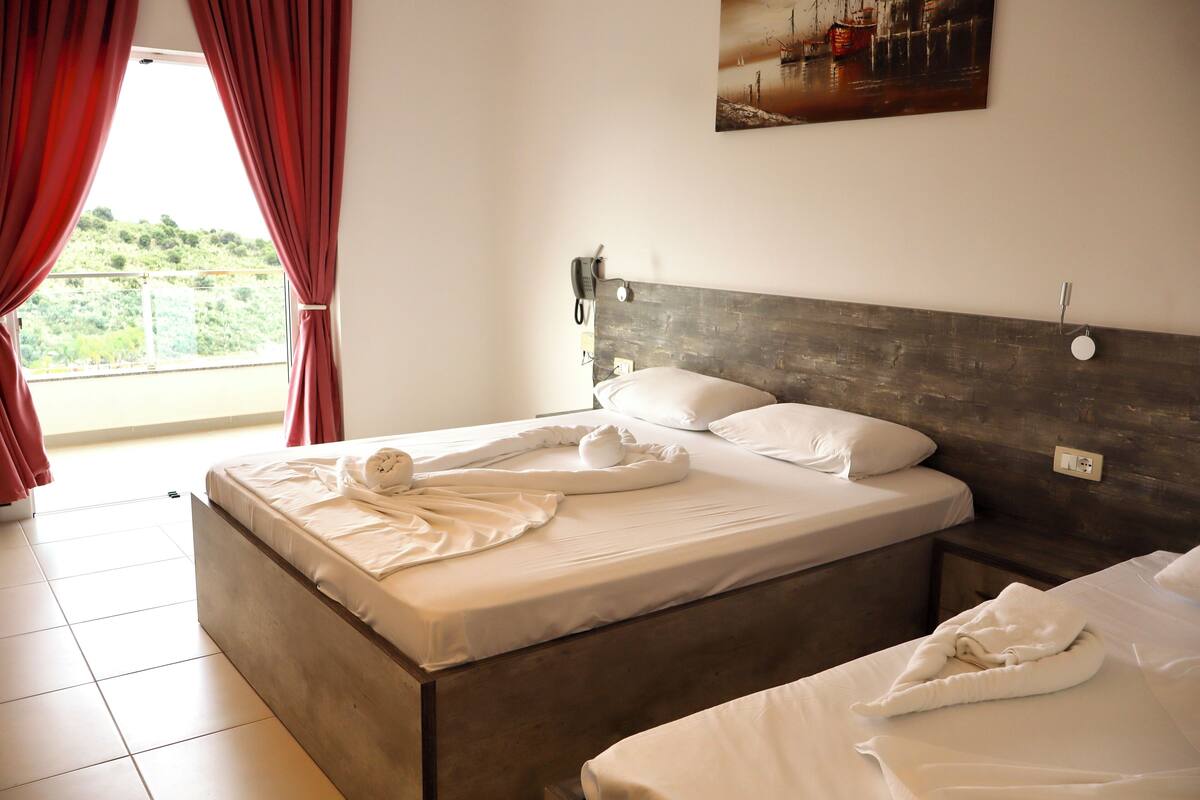A hotel room with a large bed, a wooden headboard, a painting on the wall, and a balcony with red curtains.
