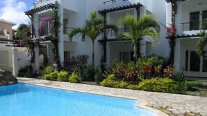 A swimming pool surrounded by a stone patio, with a white building featuring balconies and lush greenery in the background. - Aqua (Trou aux Biches)