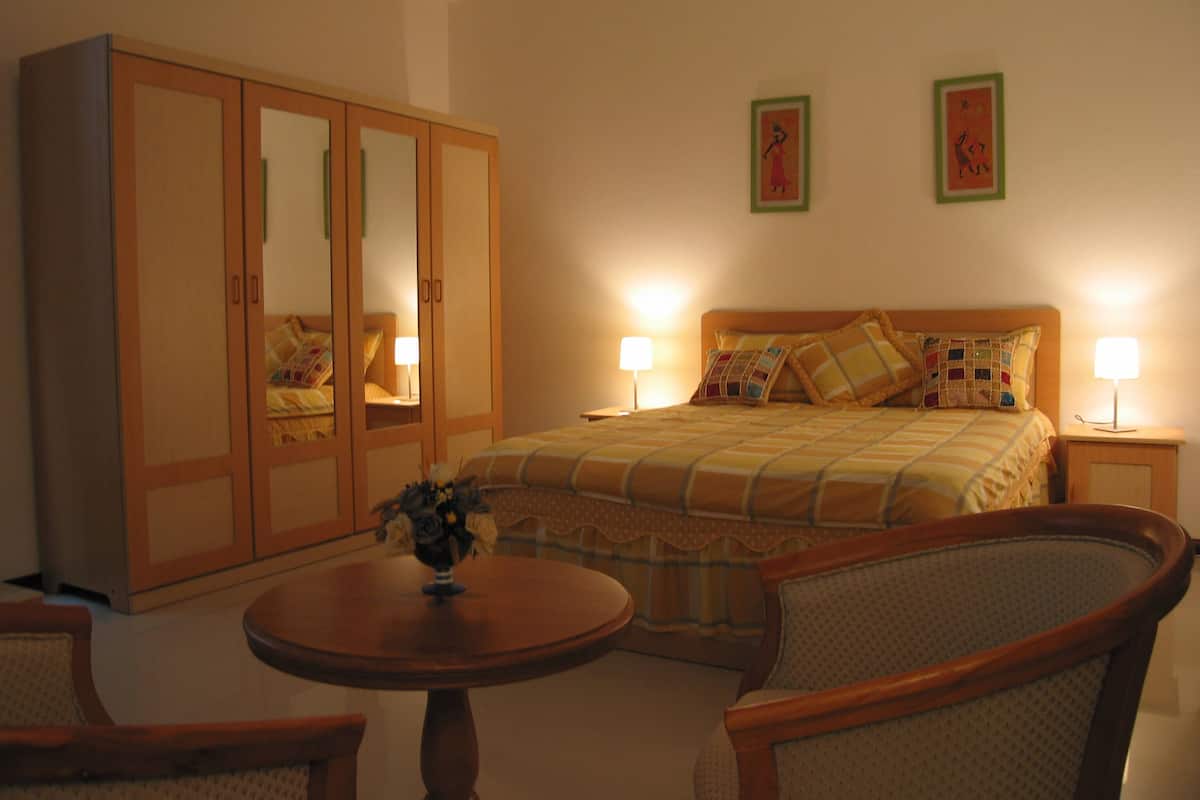A bedroom with a bed, two bedside lamps, a wooden wardrobe, a round table with a vase, and two chairs.