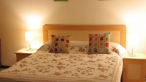 A neatly made bed with floral bedding, two decorative pillows, a bedside table with a lamp, and a small table with a vase of flowers. - Aqua (Trou aux Biches)