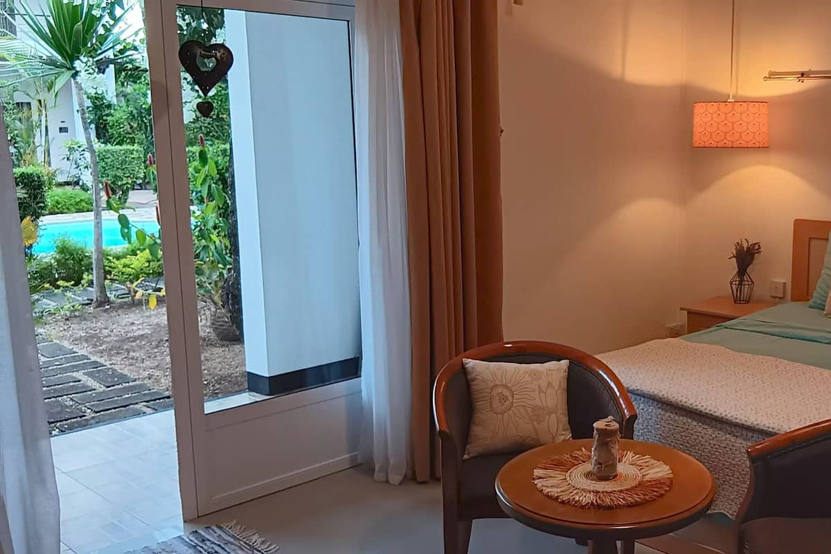 A bedroom with a bed, two chairs, a small table, a nightstand, and a view of a pool.
