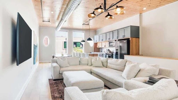 A modern living room with a sectional sofa, a flat-screen TV mounted on the wall, and a kitchen area with dark cabinets and stainless steel appliances.