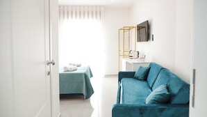 A modern hotel room with a teal sofa, a bed with a green quilt, a white bedside table, and a large window with curtains. - Gresy Boutique Apartment (Naples)