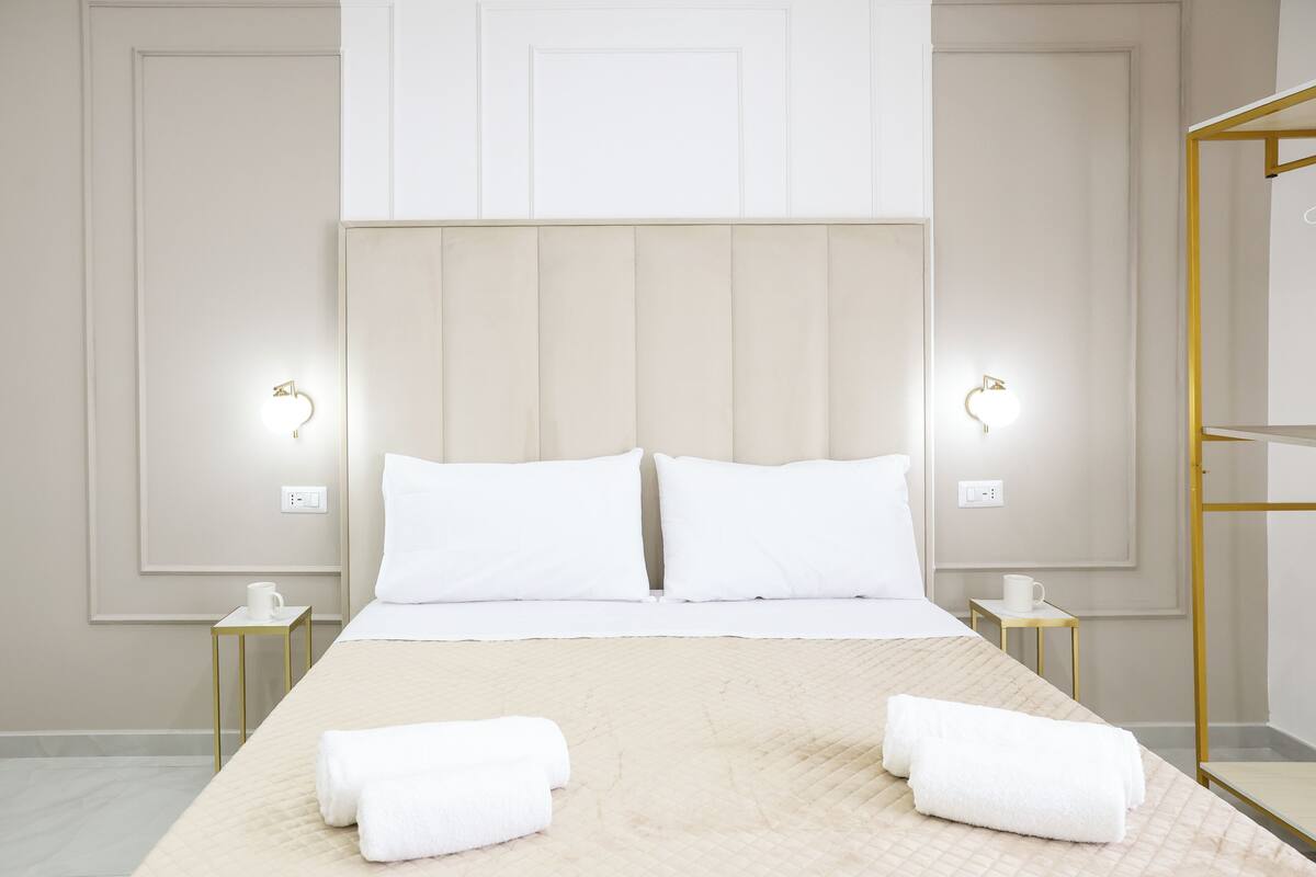 A neatly made bed with white linens, two bedside tables with lamps, and a gold-framed mirror on the right.