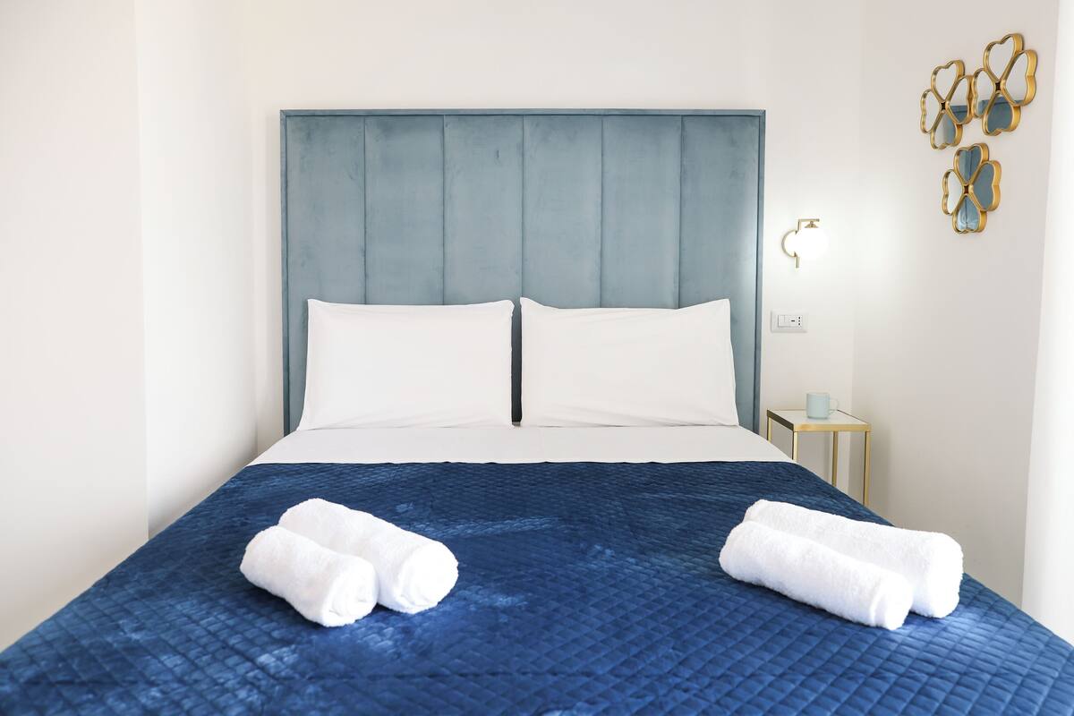 A neatly made bed with a blue quilt and two white pillows, a small bedside table with a cup, and a wall-mounted light fixture.