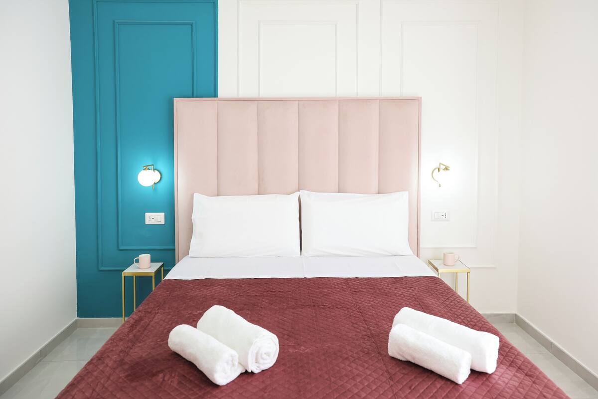 A neatly made bed with a red quilt, two white pillows, and two rolled white towels on a teal and white wall with a light switch and a small table with a pink mug.
