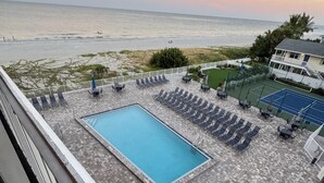 A rooftop pool area with a view of the ocean, lounge chairs, and a tennis court. - Reef Club 302 Beachfront Condo (Indian Rocks Beach)